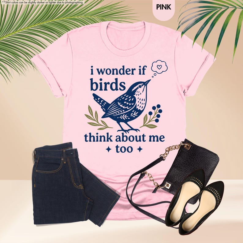 May include: A light pink short-sleeved t-shirt featuring a dark blue graphic of a bird with a thought bubble containing a heart. The text reads, "i wonder if birds think about me too." The shirt is paired with dark blue denim jeans and a black purse.