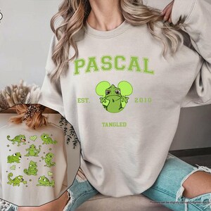 Op de afbeelding: Een zandkleurige sweatshirt met de naam "PASCAL" in groene blokletters, samen met een groen cartoonkameleonontwerp. De sweatshirt bevat ook de tekst "EST. 2010" en "TANGLED". Een kleinere afbeelding van een vergelijkbaar ontwerp is zichtbaar.