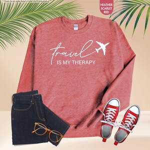 Pu&ograve; includere: Una felpa rosso scarlatto m&eacute;lange con la scritta "Travel is my Therapy" e un'immagine di un aereo bianco. L'immagine include anche un paio di jeans blu scuro, scarpe da ginnastica rosse e occhiali.