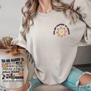 May include: A light beige sweatshirt with a graphic print on the front. The graphic is a flower with the text "You are allowed to thrive" around it. The back of the sweatshirt has a graphic print with the text "You are allowed to put yourself first, rest, outgrow people, tell yourself, speak up, walk away, be proud of yourself, change, stand up for yourself".