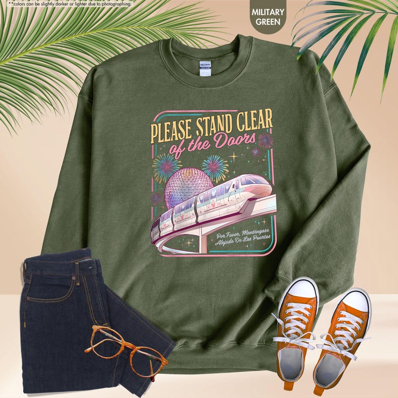 May include: Military green sweatshirt with a graphic that reads "PLEASE STAND CLEAR of the Doors". The graphic features a monorail, fireworks, and a sphere. The image also includes dark blue jeans, orange sneakers, and eyeglasses.