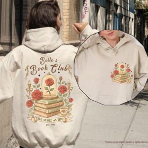Puede incluir: Sudadera con capucha color arena con un gr&aacute;fico de una rosa en una c&uacute;pula de cristal sobre libros, con el texto "Belle's Book Club" y "Tale as Old as Time". El dise&ntilde;o incluye rosas, velas y una taza de t&eacute;. La sudadera tiene un bolsillo delantero y una capucha.