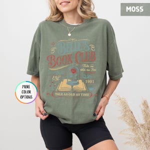 May include: Moss green t-shirt with a graphic that reads "Belle's Book Club" in a vintage style. The design includes a rose in a glass dome, an open book, and the text "Tale as old as Time" and "EST. 1991".