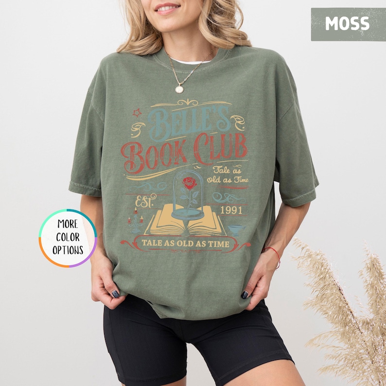May include: Moss green t-shirt with a graphic that reads "Belle's Book Club" in a vintage style. The design includes a rose in a glass dome, an open book, and the text "Tale as old as Time" and "EST. 1991".