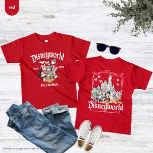 May include: Two red t-shirts with "Disneyworld" graphics. One shirt features Mickey Mouse and friends, the other shows the Disney castle. The shirts are paired with blue jeans, white sneakers, and sunglasses. The text "Est. 1971 Florida" is also visible.