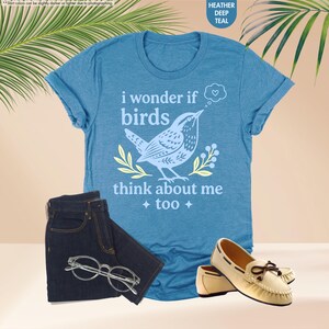 May include: A heather deep teal short-sleeve t-shirt featuring a graphic of a bird, yellow leaves, and the text "i wonder if birds think about me too" with a heart in a thought bubble. Also includes dark denim jeans, clear glasses, and beige loafers.