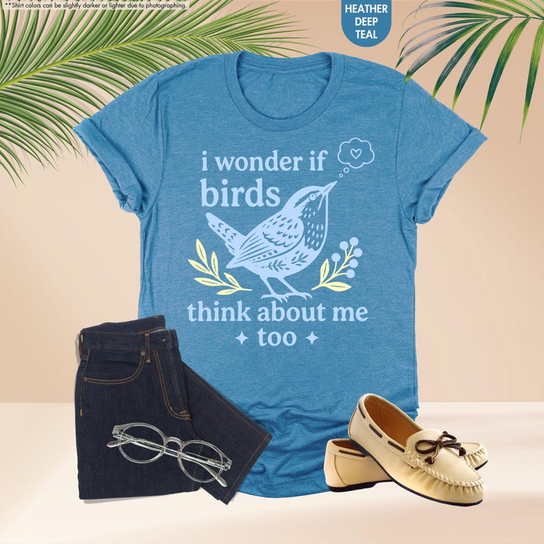 May include: A heather deep teal short-sleeve t-shirt featuring a graphic of a bird, yellow leaves, and the text "i wonder if birds think about me too" with a heart in a thought bubble. Also includes dark denim jeans, clear glasses, and beige loafers.