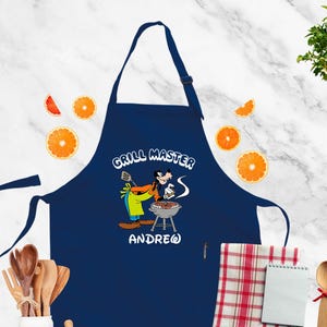 May include: Navy blue apron with a cartoon illustration of a character grilling. The apron has the text "GRILL MASTER" and the name "ANDREW". Orange slices and wooden utensils are in the background.