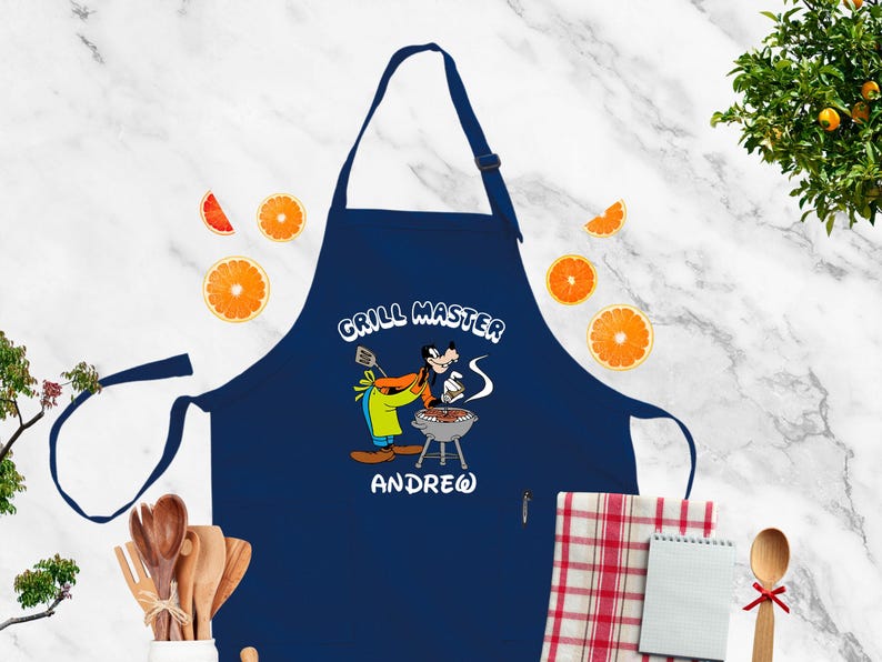 May include: Navy blue apron with a cartoon illustration of a character grilling. The apron has the text "GRILL MASTER" and the name "ANDREW". Orange slices and wooden utensils are in the background.