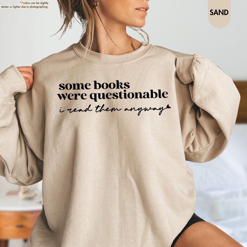 May include: A sand-colored crewneck sweatshirt featuring black text that reads some books were questionable i read them anyway. The garment has long sleeves and a ribbed neckline, cuffs, and hem, providing a standard fit for casual wear.