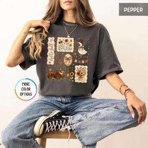 May include: A charcoal gray short-sleeve t-shirt featuring a graphic print with the text "hello fall" and various autumn-themed illustrations including a tractor, a goose wearing a hat, sunflowers, pumpkins, and a bow.