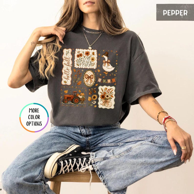 May include: A charcoal gray short-sleeve t-shirt featuring a graphic print with the text "hello fall" and various autumn-themed illustrations including a tractor, a goose wearing a hat, sunflowers, pumpkins, and a bow.