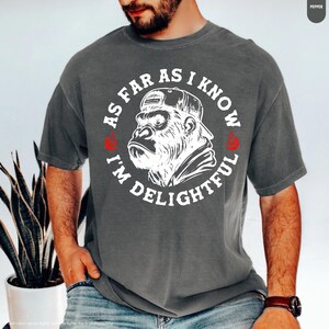 May include: Gray t-shirt with a white graphic of a gorilla wearing a backward baseball cap. The text around the gorilla reads "AS FAR AS I KNOW I'M DELIGHTFUL" in a circular design. Red accents are on the shirt.