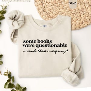 May include: A sand-colored crewneck sweatshirt featuring black text that reads, some books were questionable i read them anyway, with a small heart symbol at the end of the second line. The garment has long sleeves and a ribbed collar.
