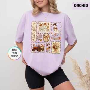 May include: A light purple t-shirt featuring a graphic print with the text hello fall. The design includes a tractor, a goose wearing a hat, floral arrangements, a bow, and autumn leaves in shades of orange, brown, and yellow on a light background.