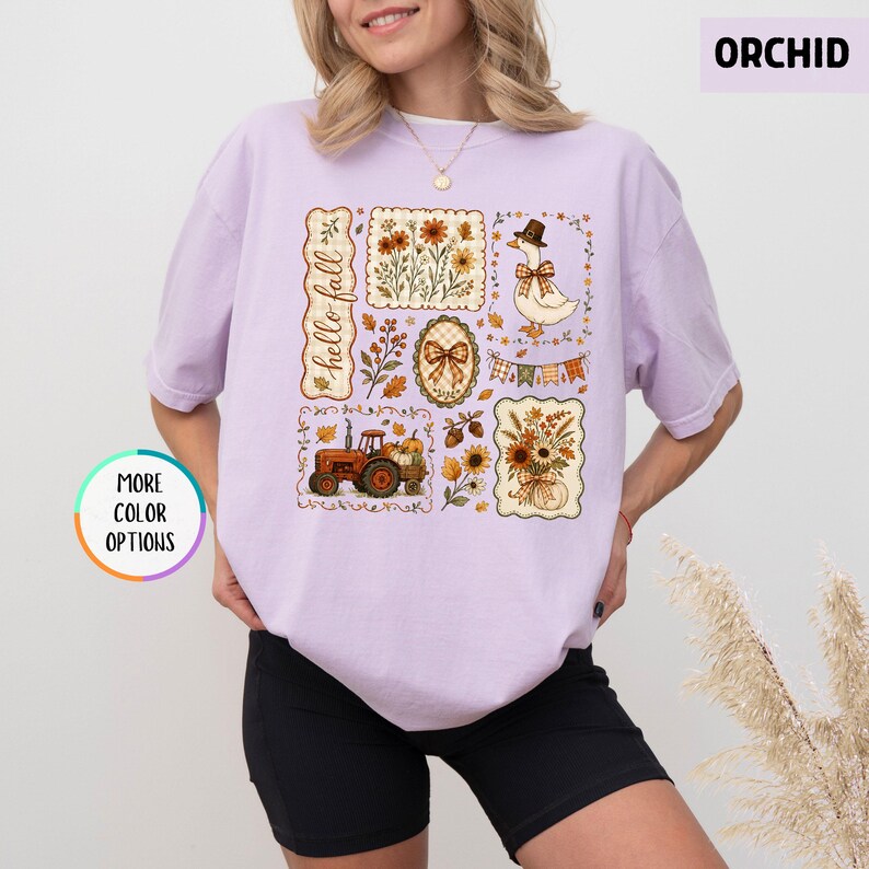 May include: A light purple t-shirt featuring a graphic print with the text hello fall. The design includes a tractor, a goose wearing a hat, floral arrangements, a bow, and autumn leaves in shades of orange, brown, and yellow on a light background.