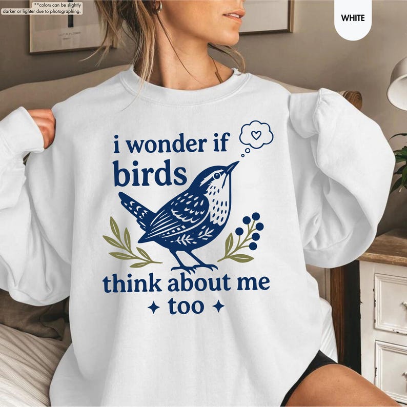 May include: A white crewneck sweatshirt featuring a dark blue graphic of a bird perched on a branch. The text reads, "i wonder if birds think about me too" with a small thought bubble containing a heart and two four-pointed star accents.
