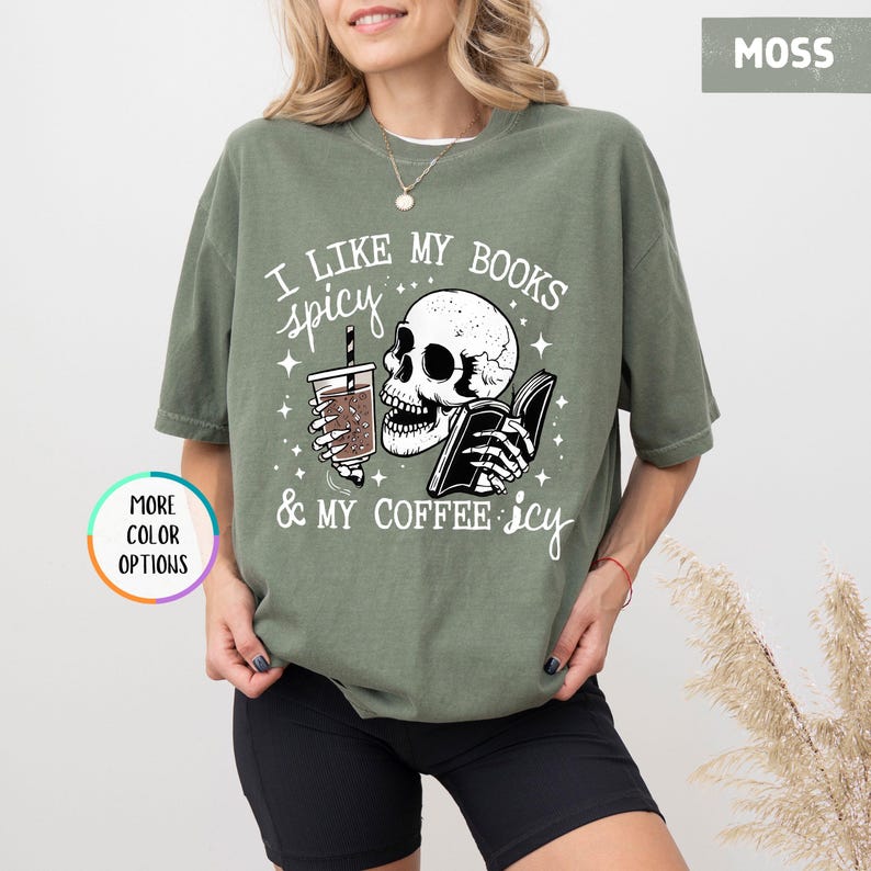 May include: A moss green t-shirt featuring a skull graphic holding a book and a coffee. The text says "I LIKE MY BOOKS spicy & MY COFFEE icy". The t-shirt has a loose, comfortable fit.