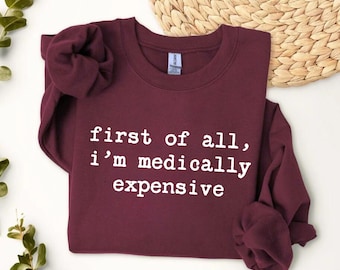 Puede incluir: Una sudadera de color granate con cuello redondo y manga larga que presenta el texto blanco "first of all, i'm medically expensive" impreso en el frente. La prenda tiene cuello, puños y dobladillo acanalados, e incluye un coletero a juego.