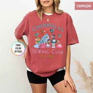 May include: A red t-shirt with a vintage-style graphic of Cinderella's Sewing Club, featuring mice characters sewing on a glass slipper. The text reads "Cinderella's Sewing Club Est. 1950".