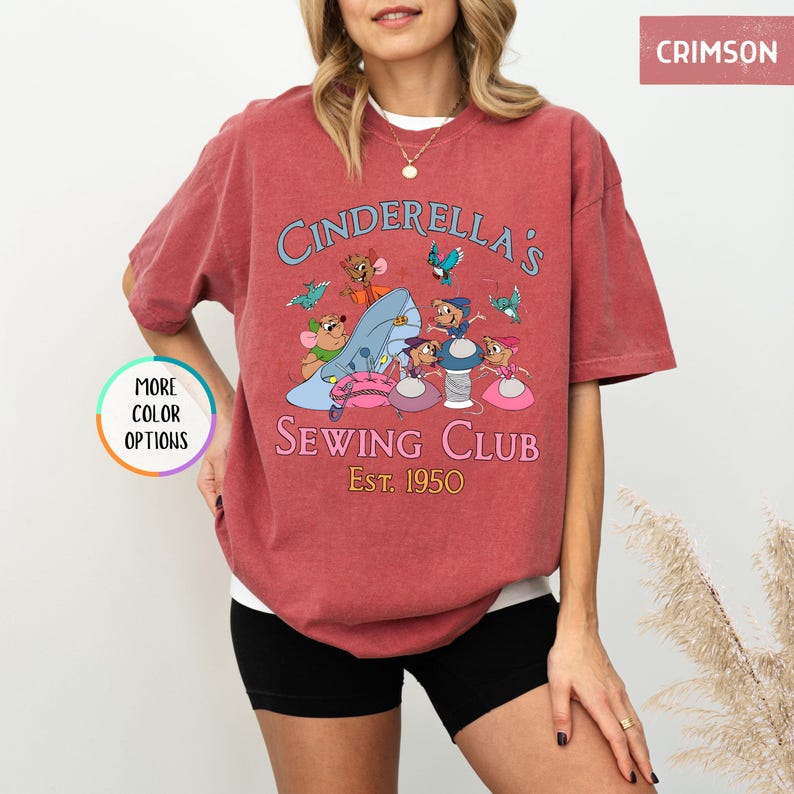May include: A red t-shirt with a vintage-style graphic of Cinderella's Sewing Club, featuring mice characters sewing on a glass slipper. The text reads "Cinderella's Sewing Club Est. 1950".