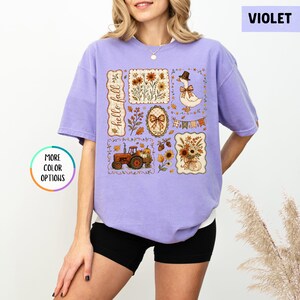 May include: A violet short-sleeve t-shirt featuring a graphic print with the text hello fall. The design includes illustrations of a tractor, a goose wearing a hat, flowers, a bow, and autumn foliage arranged in rectangular and oval frames.