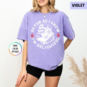 May include: A violet-colored t-shirt with a white graphic of a gorilla wearing a cap, encircled by the text "AS FAR AS I KNOW I'M DELIGHTFUL." The shirt has short sleeves and a relaxed fit. The words "MORE COLOR OPTIONS" are also visible.