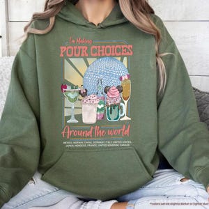 Pu&ograve; includere: Felpa con cappuccio verde militare con una grafica che dice "I'm Making Pour Choices Around the World". La grafica presenta cocktail colorati e una sfera blu. La felpa con cappuccio include un elenco di paesi.