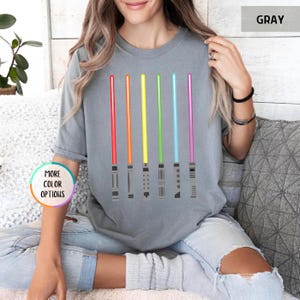 K&ouml;nnte beinhalten: Graues T-Shirt mit sechs farbigen Lichtschwertern in Rot, Orange, Gelb, Gr&uuml;n, Blau und Lila. Die Lichtschwerter sind vertikal ausgerichtet und haben detaillierte Griffe. Das Shirt hat die Worte "MORE COLOR OPTIONS" in einem Kreis.