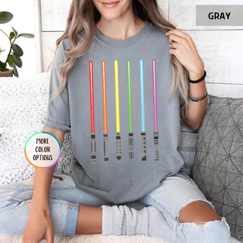 K&ouml;nnte beinhalten: Graues T-Shirt mit sechs farbigen Lichtschwertern in Rot, Orange, Gelb, Gr&uuml;n, Blau und Lila. Die Lichtschwerter sind vertikal ausgerichtet und haben detaillierte Griffe. Das Shirt hat die Worte "MORE COLOR OPTIONS" in einem Kreis.