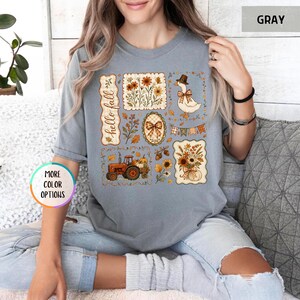 May include: A gray short-sleeve t-shirt featuring a graphic print with the text hello fall. The design includes a red tractor, a goose wearing a hat, sunflowers, pumpkins, acorns, and a plaid bow, all arranged in a collage of rectangular frames.