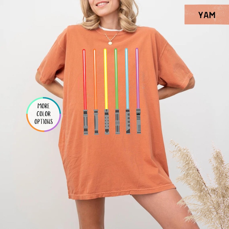 K&ouml;nnte beinhalten: Ein orangefarbenes T-Shirt mit einem Regenbogen-Lichtschwert-Design. Die Lichtschwerter sind rot, orange, gelb, gr&uuml;n, blau und lila. Das Shirt hat den Text "MORE COLOR OPTIONS" links und "YAM" oben rechts.