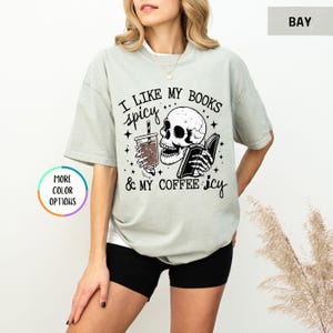 May include: A pale green t-shirt featuring the phrase "I like my books spicy & my coffee icy" alongside a skull graphic holding a book and a drink. The top has short sleeves and a loose fit.