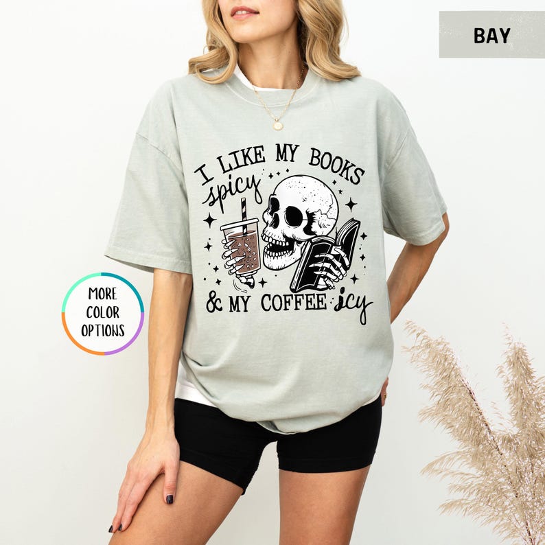 May include: A pale green t-shirt featuring the phrase "I like my books spicy & my coffee icy" alongside a skull graphic holding a book and a drink. The top has short sleeves and a loose fit.