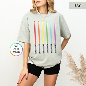 K&ouml;nnte beinhalten: Hellgr&uuml;nes T-Shirt mit einem Regenbogen-Lichtschwert-Design. Die Lichtschwerter sind rot, orange, gelb, gr&uuml;n, blau und lila. Das Shirt hat eine lockere Passform und wird mit schwarzen Shorts getragen.