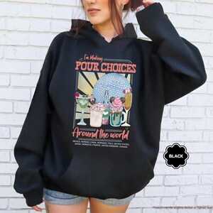 Pu&ograve; includere: Felpa con cappuccio nera con una grafica che recita "I'm Making POUR CHOICES Around the world". La grafica presenta illustrazioni di cocktail e un globo. La felpa con cappuccio &egrave; nera con cappuccio con coulisse e tasca frontale.
