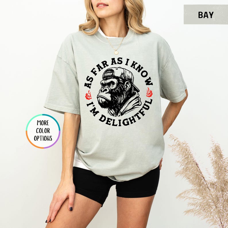 May include: A light green t-shirt with a graphic of a gorilla wearing a cap. The text around the gorilla reads "AS FAR AS I KNOW I'M DELIGHTFUL". The shirt has a relaxed fit and short sleeves. The design is in black and red.