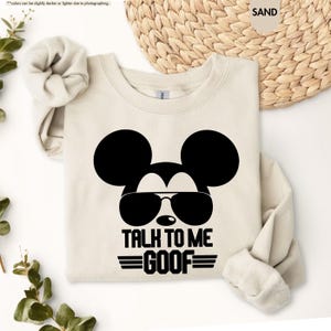 May include: A sand-coloured sweatshirt with a black graphic of Mickey Mouse wearing sunglasses. The text "TALK TO ME GOOF" is below the graphic in black. The sweatshirt has long sleeves and a crew neck.