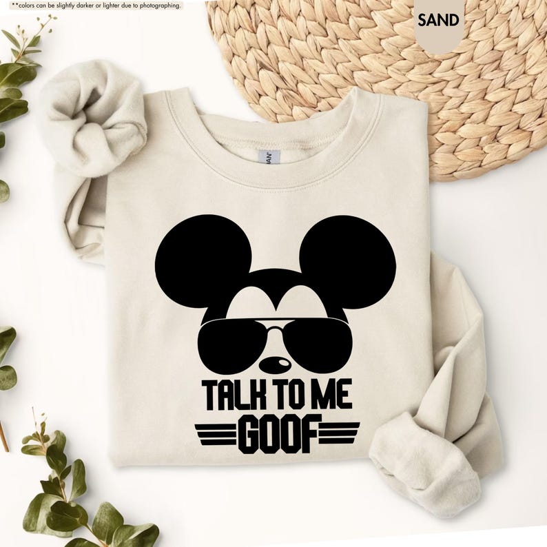May include: A sand-coloured sweatshirt with a black graphic of Mickey Mouse wearing sunglasses. The text "TALK TO ME GOOF" is below the graphic in black. The sweatshirt has long sleeves and a crew neck.