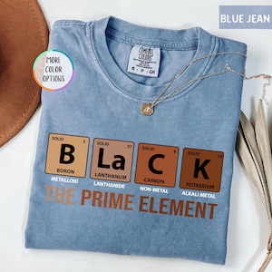 May include: A light blue t-shirt with the word "BLACK" spelled out using periodic table elements. The phrase "THE PRIME ELEMENT" is printed below. A gold necklace is draped across the shirt.