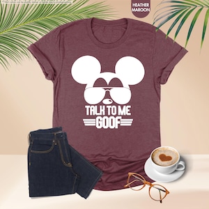 May include: Maroon heather t-shirt featuring a white Mickey Mouse graphic wearing sunglasses and the text "TALK TO ME GOOF". Dark blue denim jeans are folded beside the shirt. A cup of coffee and glasses are also in the image.
