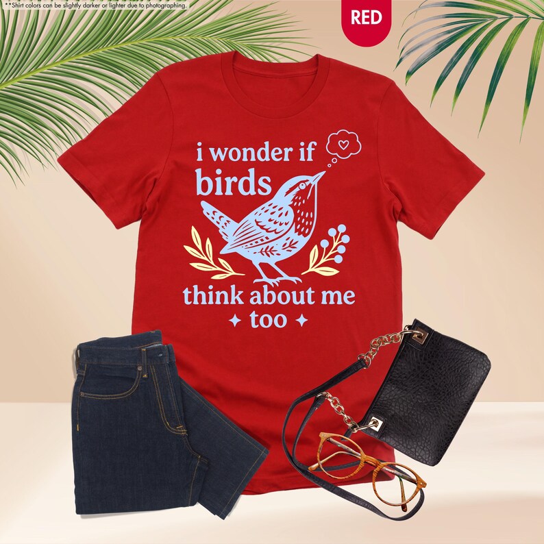 May include: A red short-sleeved t-shirt featuring a light blue graphic of a bird, yellow sprigs, and the text "i wonder if birds think about me too" with a heart in a thought bubble. The shirt is paired with dark blue jeans, a black bag, and glasses.