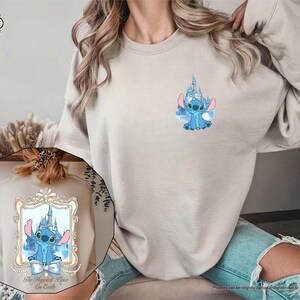 May include: Sand-colored sweatshirt featuring a blue cartoon character with a castle design. The sweatshirt has a crew neck and long sleeves. The design includes the text "The Happiest Place on Earth" in a decorative frame.