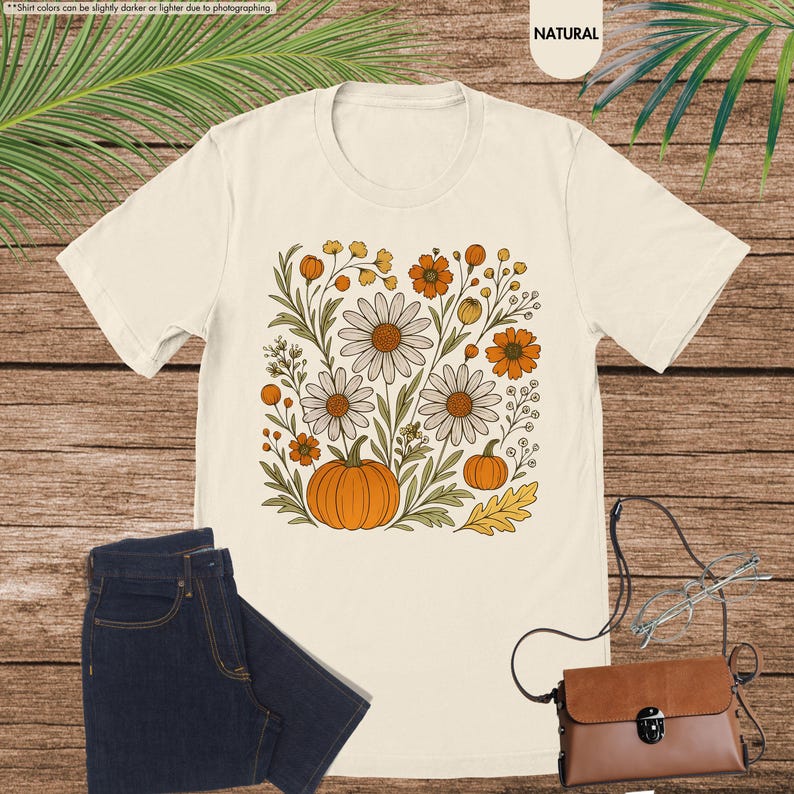 May include: Cream-coloured t-shirt with a floral design featuring daisies, orange flowers, and pumpkins. The design is centered on the shirt. The shirt is paired with dark blue jeans and a brown purse with glasses.