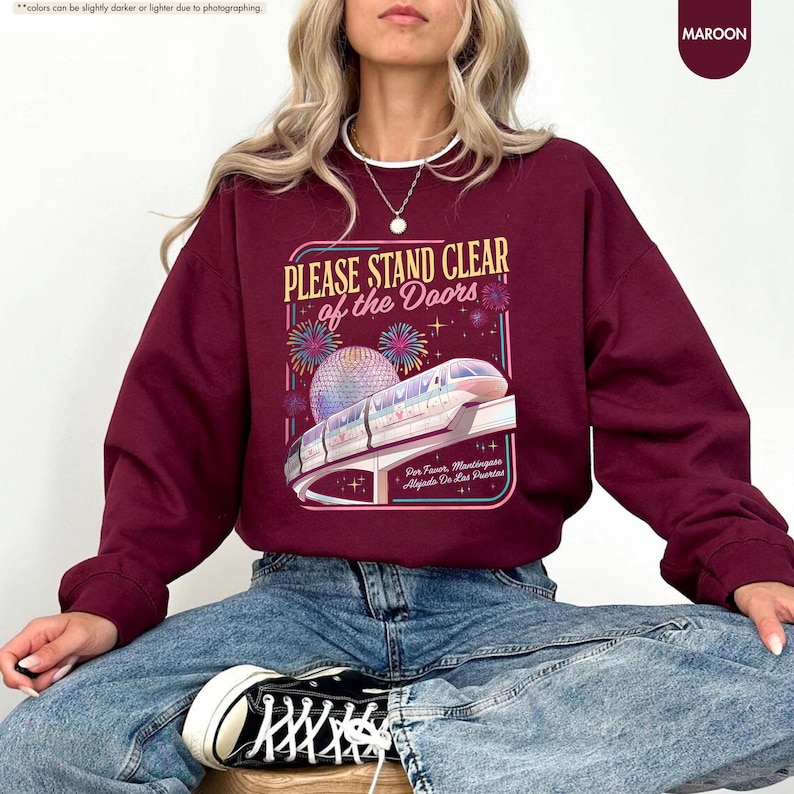 May include: Maroon sweatshirt with the text "PLEASE STAND CLEAR of the Doors" and a graphic of a monorail and fireworks. The sweatshirt is a crew neck style and is paired with blue jeans and black sneakers.