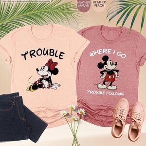 May include: Two heather t-shirts, one peach and one mauve, featuring cartoon characters. The peach shirt has "TROUBLE" and a Minnie Mouse graphic. The mauve shirt says "WHERE I GO TROUBLE FOLLOWS" with a Mickey Mouse graphic. Also shown are jeans and shoes.