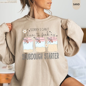 May include: A tan sweatshirt with a graphic of three glass jars with pink lids and the text "SORRY I CANT i have to feed my SOURDOUGH STARTER".