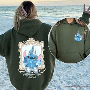 May include: Olive green hoodie featuring a cartoon image of Stitch in front of a castle, framed with a decorative border and the text "The Happiest Place On Earth." A smaller Stitch graphic is on the chest. The hoodie has a front pocket.