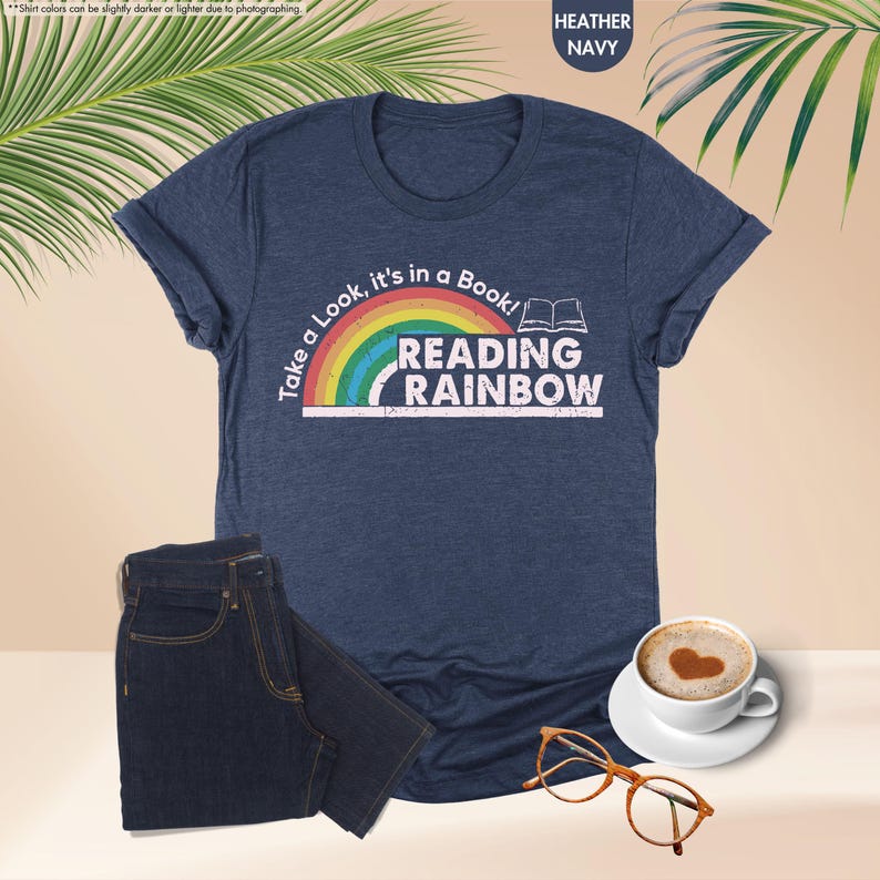 May include: Heather navy blue t-shirt with the text "Take a Look, it's in a Book! READING RAINBOW" above a rainbow graphic. A pair of dark blue jeans, a cup of coffee, and eyeglasses are also in the image.