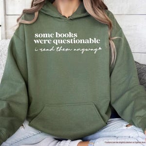 May include: A military green hooded sweatshirt featuring the white printed text some books were questionable i read them anyway. The garment includes a front kangaroo pocket, long sleeves with ribbed cuffs, and a drawstring hood in a matching color.